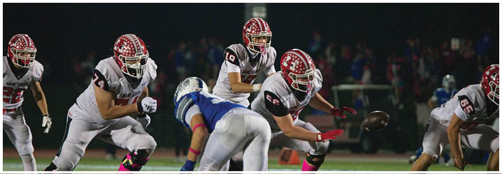 People in football uniforms are actively engaged in a play on a nighttime field with one player in white and red running while others in white and blue are attempting to tackle. Description generated by AI