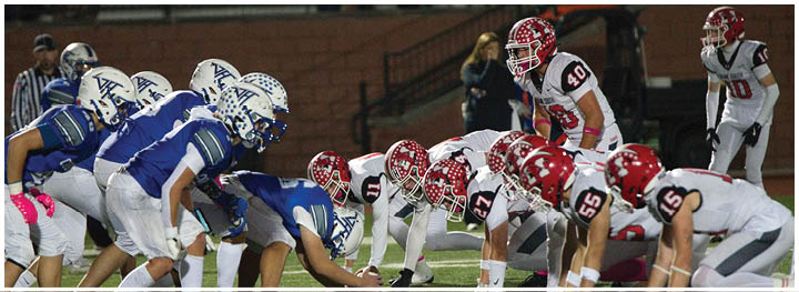 Two opposing American football teams in blue and white and red and white uniforms are lined up facing each other on a field, ready for a play. Description generated by AI