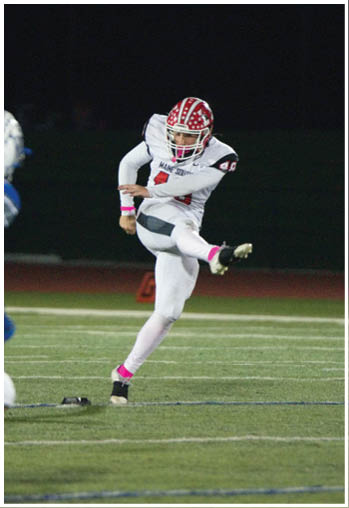 A person in a white football uniform with a red helmet is kicking a football on a field while another person in a blue uniform watches. Description generated by AI