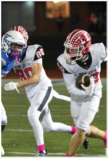 Two people in white football uniforms with red helmets are running on a field, with one holding a football and wearing pink socks and cleats. Description generated by AI