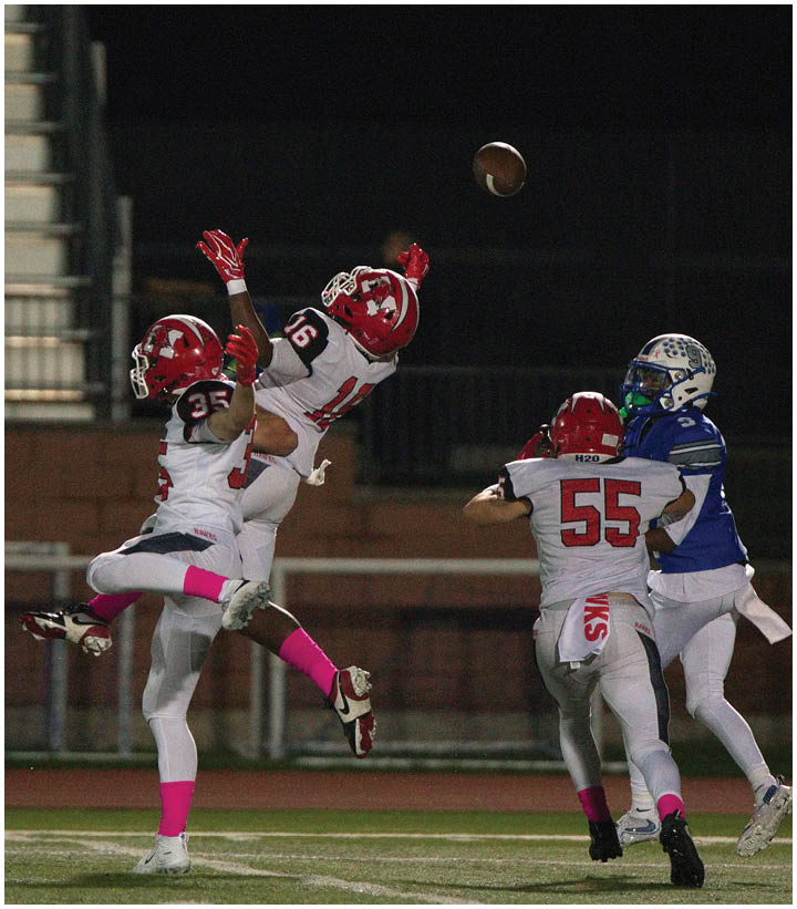 Three people in white and red football uniforms are jumping to catch a football while a person in a blue and white uniform approaches on a field at night. Description generated by AI