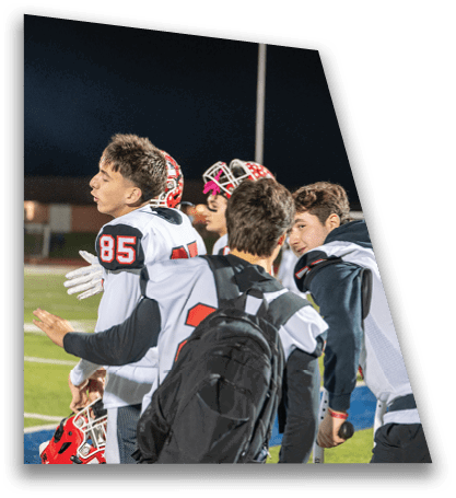 People wearing football uniforms and helmets are gathered on a field at night, with one person carrying a backpack and another holding a microphone. Description generated by AI