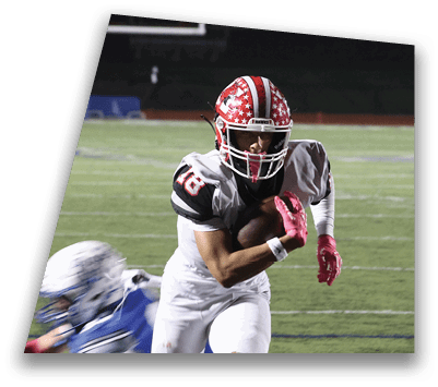 Two people in football uniforms are on a field at night with one running while holding the ball and the other following behind. Description generated by AI