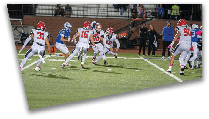 People in football uniforms are actively playing on a field with spectators in the stands during a game at dusk. Description generated by AI
