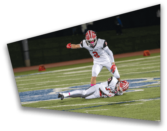 Two people in football uniforms are on a field with one person lying on the ground and the other standing nearby. Description generated by AI