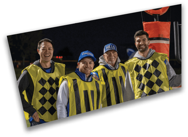 Three adults wearing yellow vests and smiling stand outdoors at night, with one wearing a blue cap that says “Cougars" and another wearing a blue cap that says "HILLS." Description generated by AI