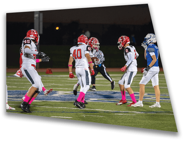 Four people in football uniforms with red helmets and pink socks are on a field facing one person in a blue and white uniform and helmet, with a referee in the background. Description generated by AI