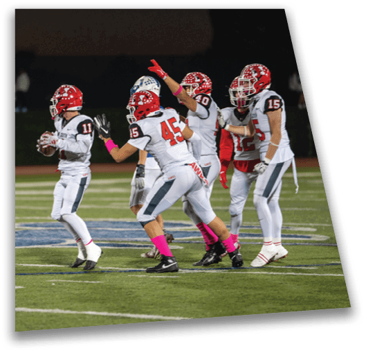 People in football uniforms are on a field with one person holding a football and others appearing to block or prepare for a play during a game. Description generated by AI