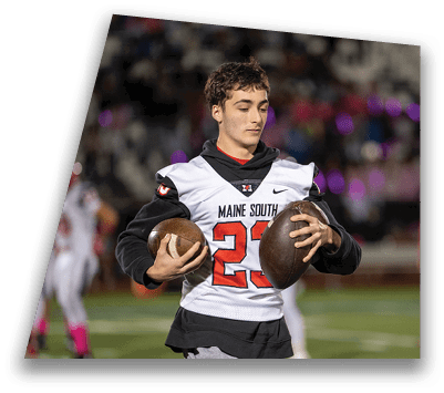A person wearing a Maine South football jersey and gray sweatpants is walking on a football field holding two footballs with players and spectators blurred in the background. Description generated by AI