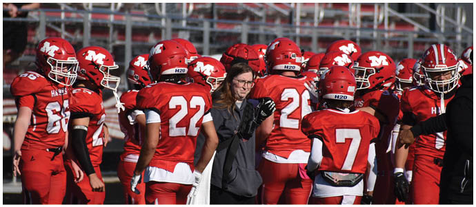 A group of people wearing red football uniforms and helmets are gathered on a field with one person in casual clothing holding a football. Description generated by AI