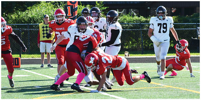 Players in red and white football uniforms are engaged in a tackle on a green field while other players and a person in casual clothing observe in the background. Description generated by AI