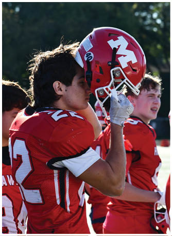 Three people in red football uniforms stand outdoors, with one holding a red helmet featuring a white “M" logo. Description generated by AI