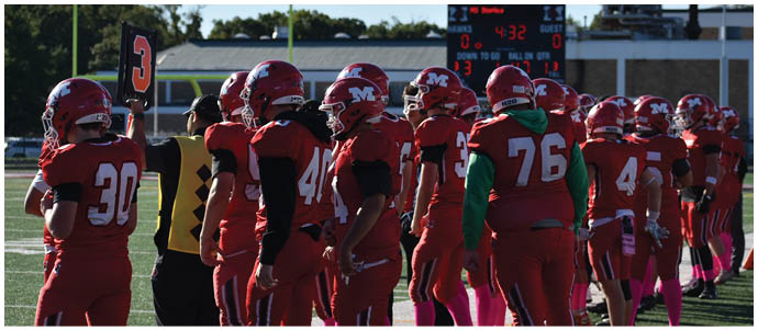 A group of people wearing red football uniforms and helmets stand on a field near a person holding a substitution board. Description generated by AI