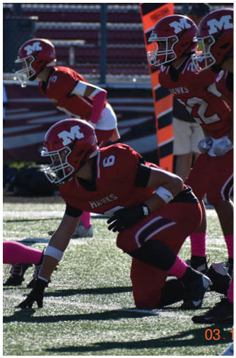 A person in a red football uniform with a helmet and gloves is crouched on a field, preparing for a play. Description generated by AI