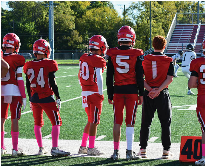 A group of people wearing red football uniforms and pink socks stand on the sidelines of a football field next to a person in black pants and a red shirt. Description generated by AI