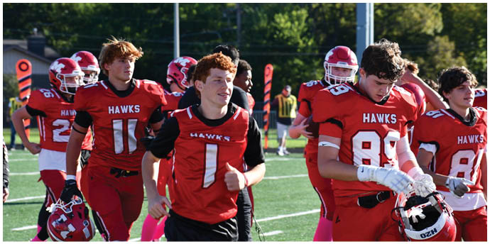Several people wearing red football uniforms with “HAWKS" on the front are walking on a green field, with one holding a helmet. Description generated by AI