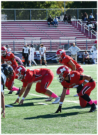 Three people in red football uniforms and helmets are crouched in a ready position on a green field with others and empty bleachers in the background. Description generated by AI