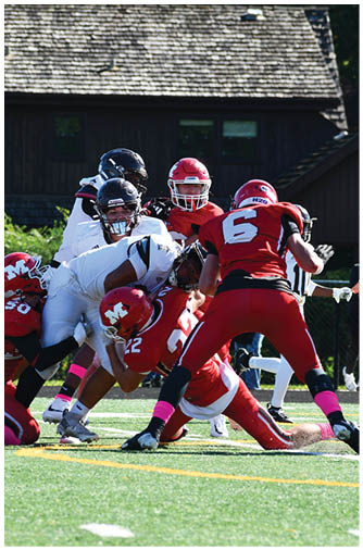 A group of football players in red and white uniforms are engaged in a play on a sunny field with a building and spectators in the background. Description generated by AI