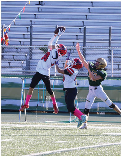 Three people in football uniforms are actively reaching for a football in midair on a field with empty bleachers in the background. Description generated by AI
