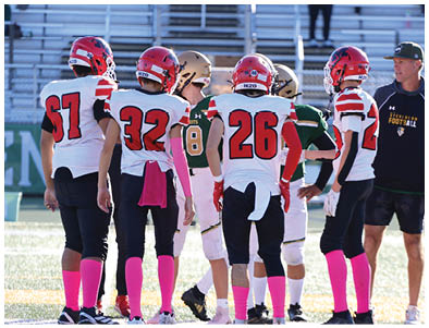 Five people wearing football uniforms with red helmets and pink socks stand on a field facing two others in green and gold uniforms during a game. Description generated by AI