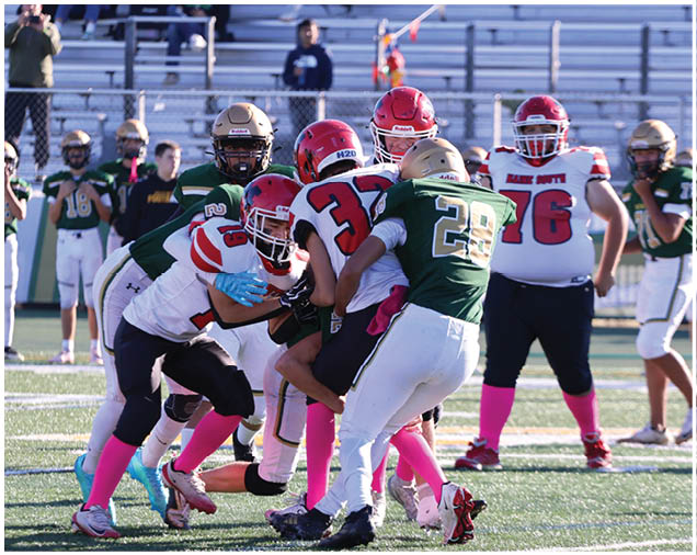 Several people wearing football uniforms with helmets and pink socks are engaged in a tackle on a football field with others watching in the background. Description generated by AI