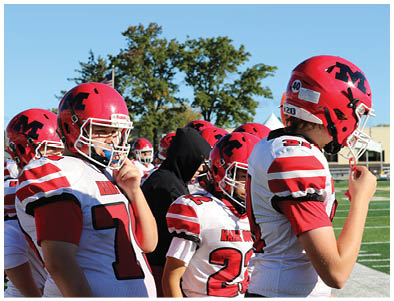 Several people wearing red and white football uniforms and helmets stand together on a sports field under a clear blue sky. Description generated by AI