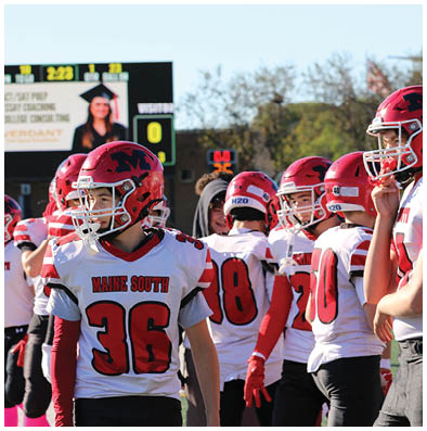 A group of people wearing red helmets and white football jerseys with red numbers are standing on a field with a scoreboard visible in the background. Description generated by AI