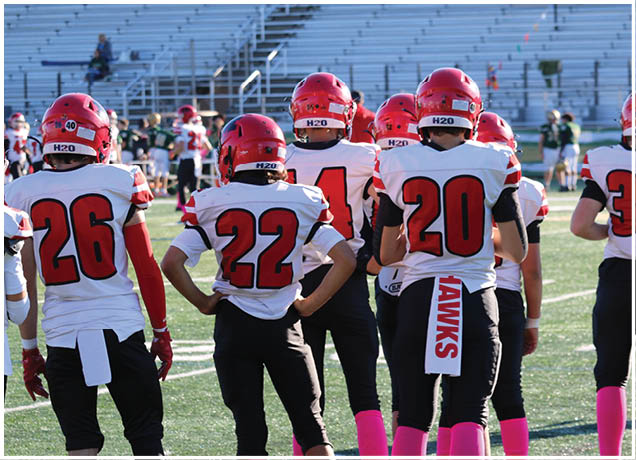 A group of people wearing red helmets, white jerseys with large red numbers, and black pants are standing on a sports field, likely preparing for a football game. Description generated by AI