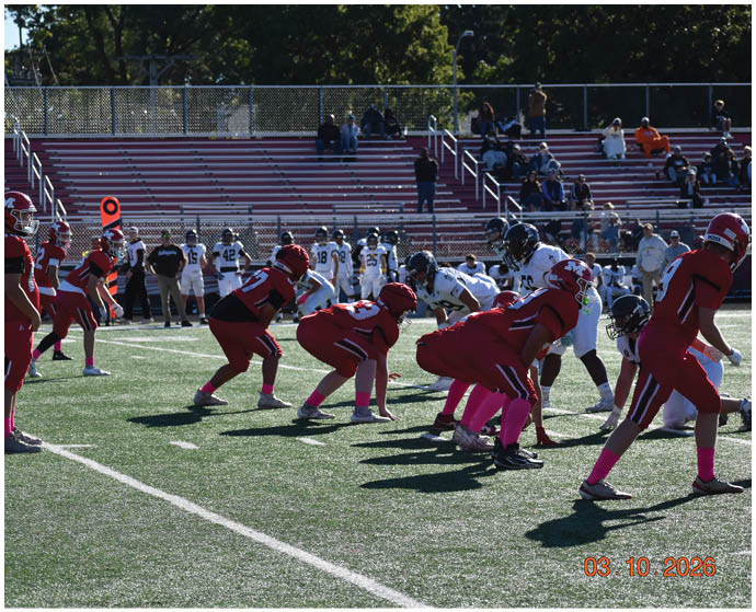 A group of people in red sports uniforms with pink socks are lined up on a football field facing a team in white uniforms during a game with spectators in the background. Description generated by AI