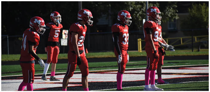 Five people wearing red football uniforms and pink socks stand on a football field, appearing to prepare for a play. Description generated by AI