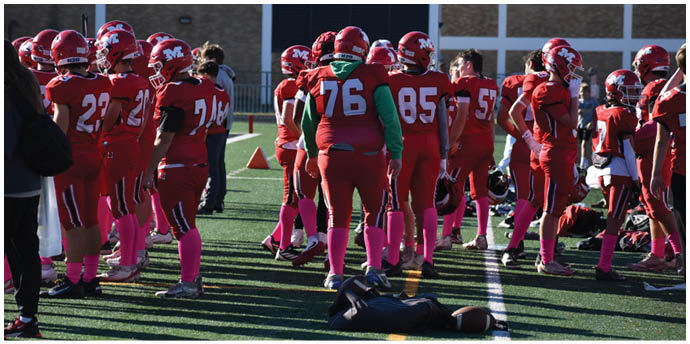 A group of people wearing red sports uniforms and pink socks stand on a field while one person lies on the ground near a football. Description generated by AI
