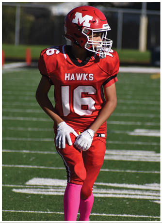 A person wearing a red football uniform with the number 16 and the word “HAWKS" on the jersey stands on a football field with green turf and white yard lines. Description generated by AI