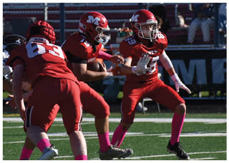 Three people in red football uniforms are on a field with one holding a football and another preparing to receive it. Description generated by AI