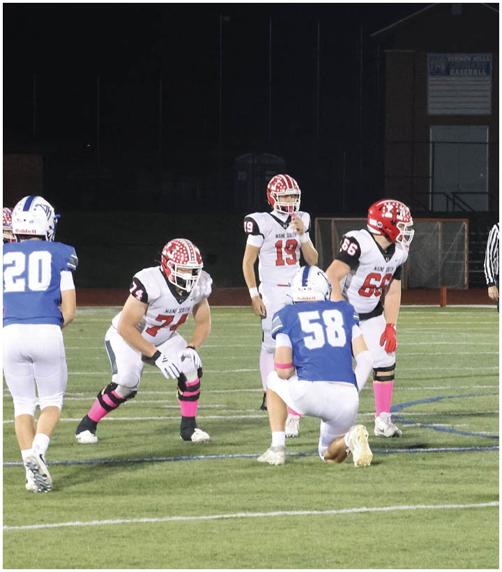 A group of football players in white and red uniforms and one player in a blue uniform are positioned on a field at night with a referee standing behind them. Description generated by AI