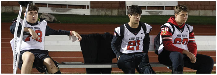 Two people wearing Maine South football jerseys are sitting on a bench at a sports field, appearing relaxed and watching the game. Description generated by AI