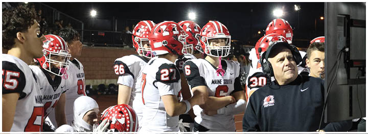 A group of people wearing football helmets and uniforms stand together on a field while a person in a headset and black jacket gestures and looks to the side. Description generated by AI