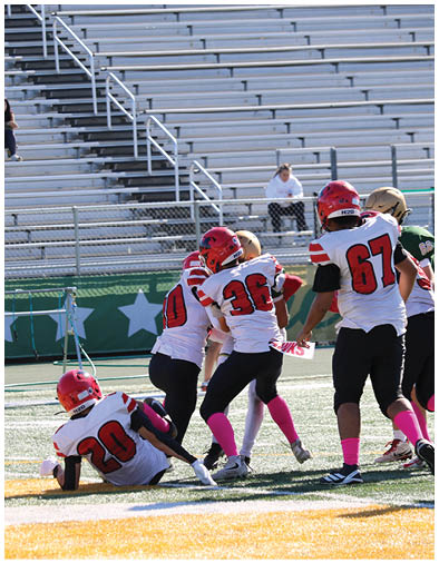 Four people in football uniforms with red helmets and pink socks are engaged in a play on a football field with empty bleachers in the background. Description generated by AI