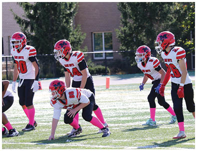 Four children in football uniforms with red helmets and pink socks are positioned on a grassy field preparing for a play during a game. Description generated by AI