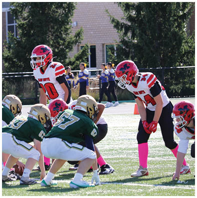 People in football uniforms are lined up on a field preparing for a play during a game with a building and trees in the background. Description generated by AI