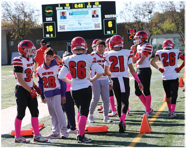 People wearing football uniforms with pink socks and accessories are standing and talking on a football field near an orange cone with a scoreboard visible in the background. Description generated by AI