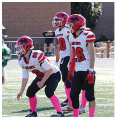 Three people in red helmets and white football jerseys with pink socks are positioned on a football field while another person in a green and gold uniform stands nearby. Description generated by AI