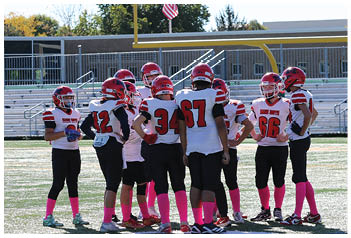 A group of people wearing red helmets and white sports jerseys with pink socks are standing together on a football field near bleachers and a goalpost with an American flag in the background. Description generated by AI