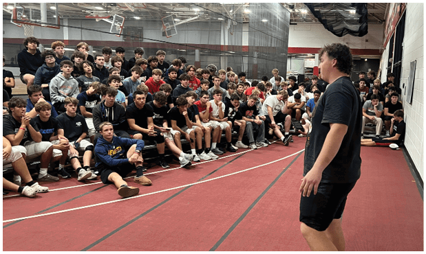 A person in athletic clothing stands on a red indoor track facing a large group of seated people who appear to be listening attentively. Description generated by AI