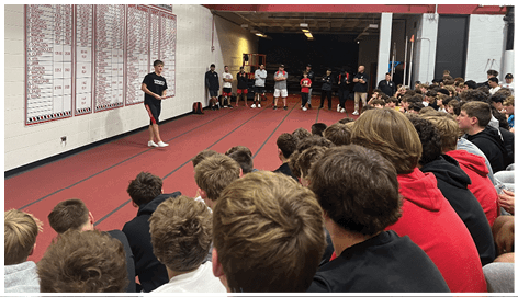 A person stands on an indoor track speaking to a large seated audience of people who are attentively watching. Description generated by AI