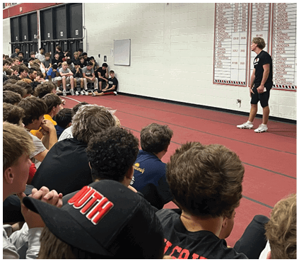 A large group of people sitting closely together on bleachers in an indoor gymnasium, attentively facing toward the right side of the image. Description generated by AI