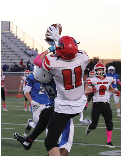 A person wearing a red helmet and white jersey with the number 11 is catching a football while other players in red and blue uniforms watch on a football field at sunset. Description generated by AI