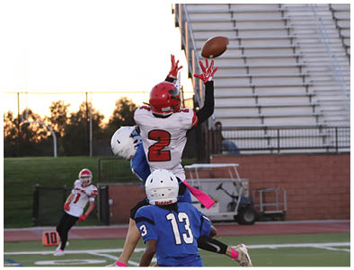 Two people in football uniforms are jumping to catch a ball while another person in a blue uniform watches closely on a football field during sunset. Description generated by AI