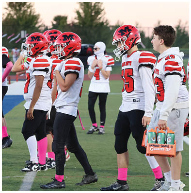 Several people wearing red and white football uniforms stand on a field with one person holding a container of sports drinks. Description generated by AI