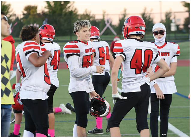 A group of people wearing white and red football uniforms are standing on a field, with one person holding a helmet and another wearing a white head covering and sunglasses. Description generated by AI