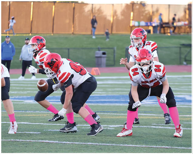Four people in red helmets and white jerseys with pink socks are positioned on a football field, with one person holding a football ready to snap it. Description generated by AI